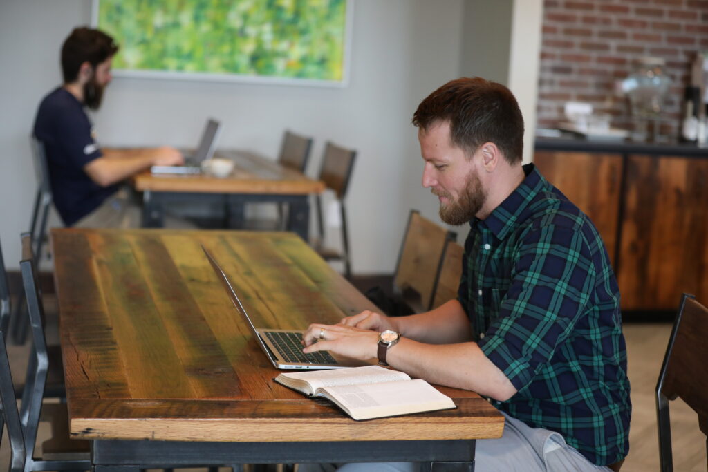 A man with a beard and plaid shirt works on a laptop at a wooden table with an open book, possibly handling BSOL project management tasks. Another person is in the background, also using a laptop in the casual café or workspace setting.