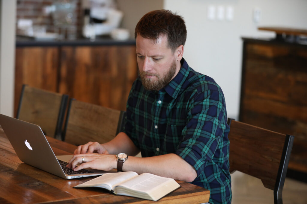 A man with a beard, wearing a green plaid shirt, sits at a wooden table typing on a laptop with an open book in front of him, focused on his Associate of Science in Professional Studies. The background shows a blurred indoor setting.
