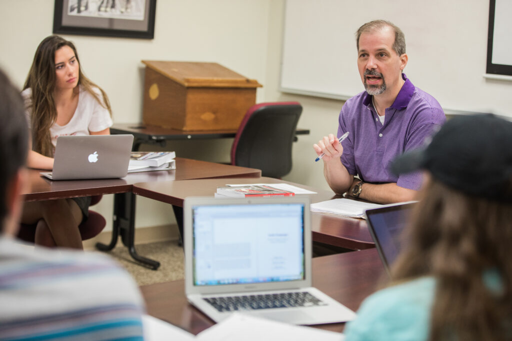A man in a purple shirt speaks to a small group in a classroom, discussing International Relations. Several people are seated around tables with laptops open, listening. A whiteboard and framed picture are visible in the background.