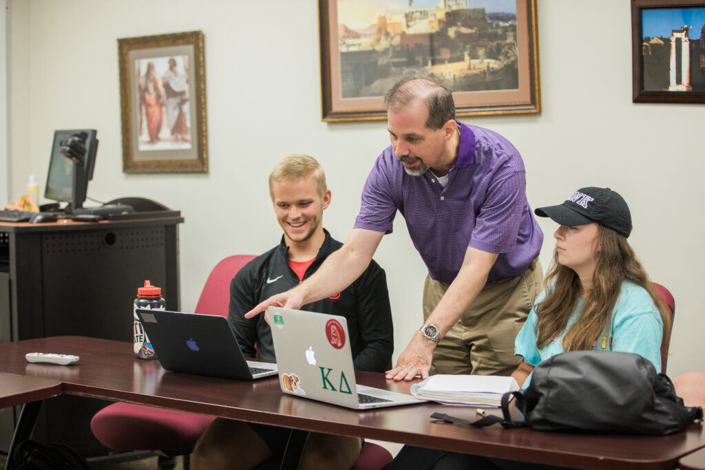 A man in a purple shirt stands and points at a laptop screen while two students studying International Affairs sit at a table with their laptops and books in a classroom. Framed art hangs on the wall behind them.