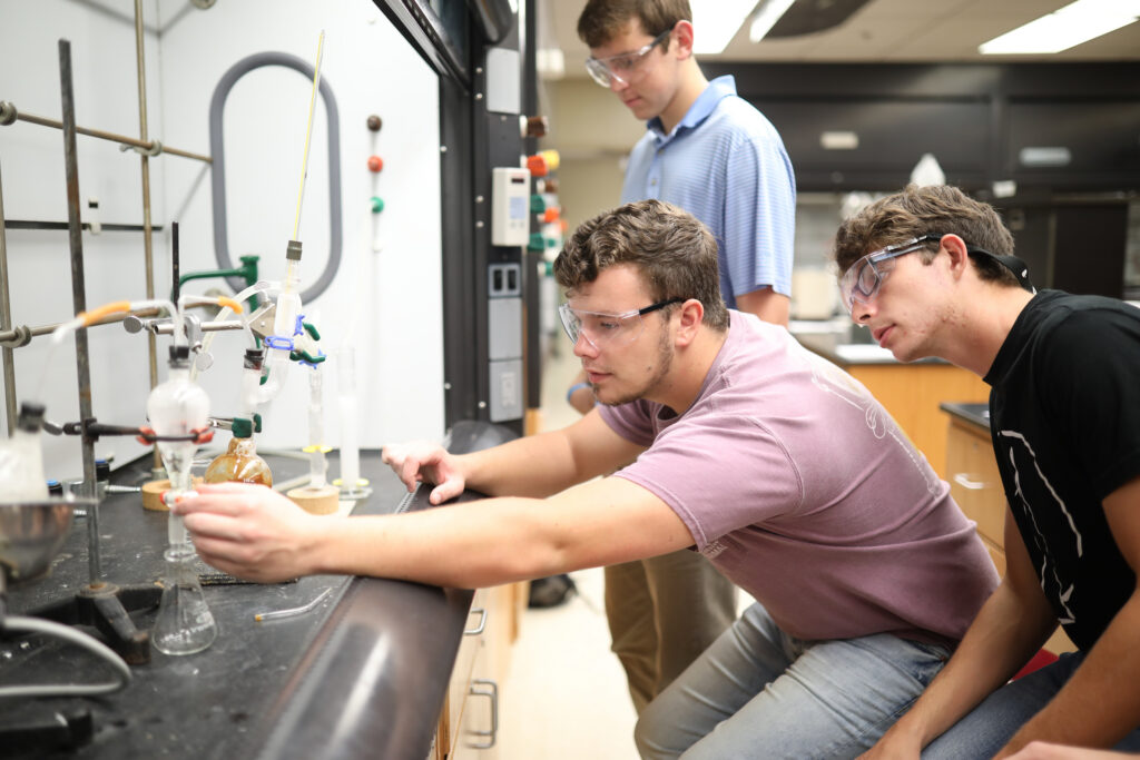 Three young men in safety glasses work at a laboratory bench with scientific equipment.