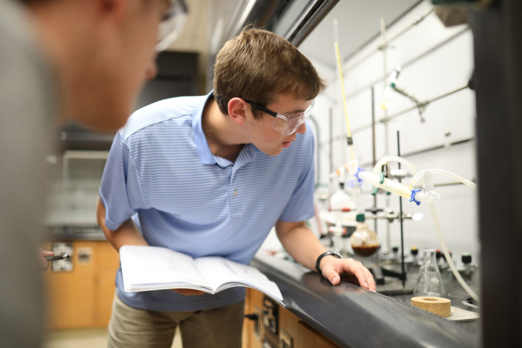 A person wearing safety goggles and a light blue striped shirt looks closely at a chemistry experiment setup in a laboratory while holding an open notebook. Glassware and lab equipment are visible on the counter.