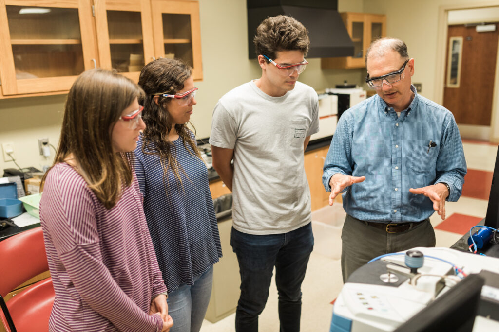 A man in safety glasses explains a scientific instrument to three students, also wearing safety glasses, in a medical laboratory with wooden cabinets and equipment.