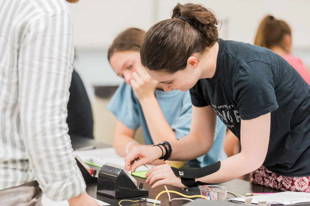 A student uses a tape dispenser at a classroom table while another student watches. Papers, wires, and classroom supplies are on the table. Two more students are visible in the background.