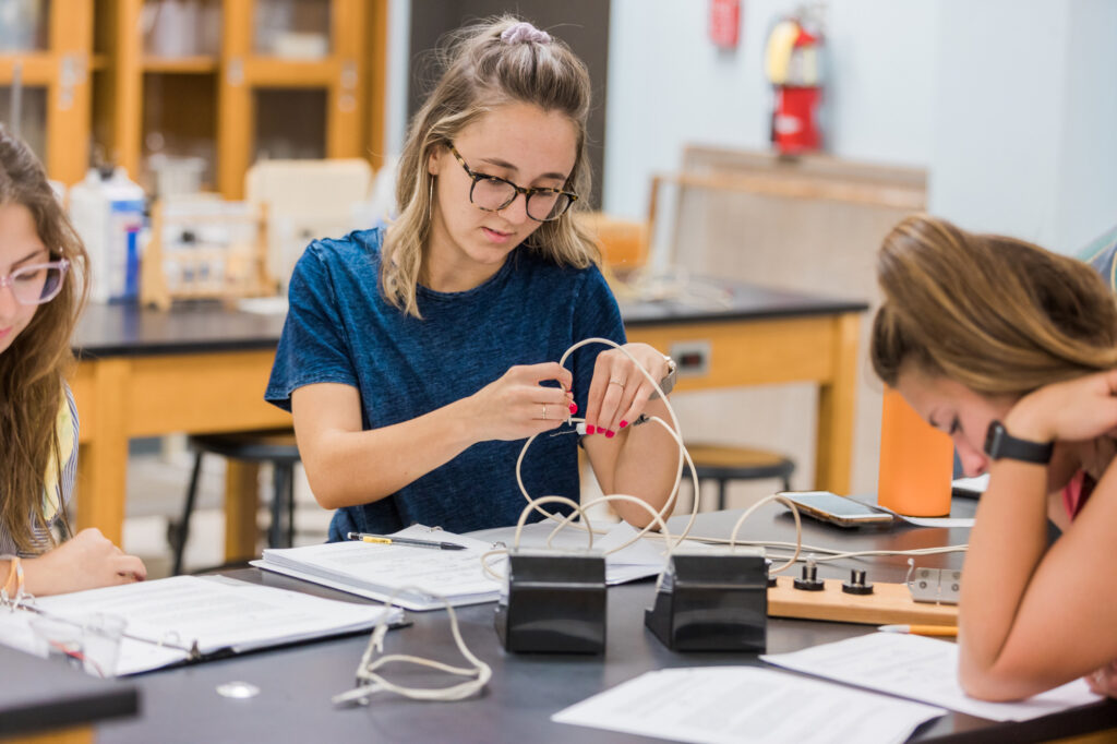 Three students sit at a lab table, working with electronic equipment and wires. One student is adjusting wires, while another writes in a notebook. Lab materials and papers are spread on the table.