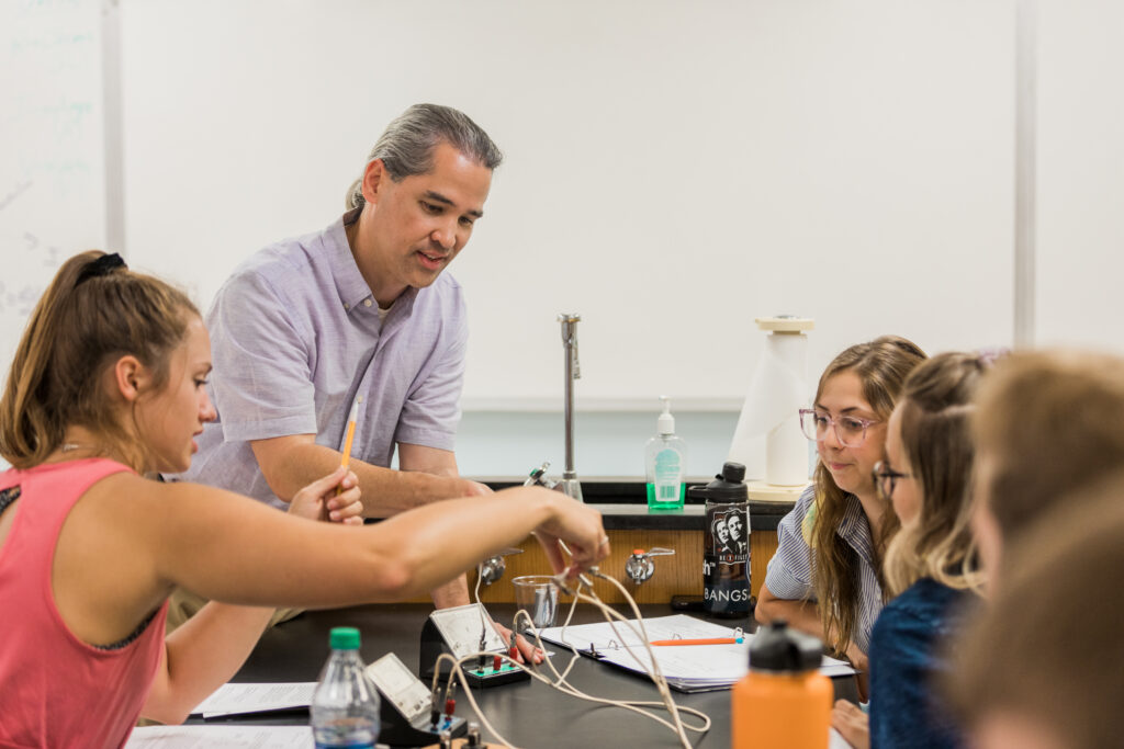 A teacher stands at a lab table assisting a group of students working on a science experiment. Lab equipment, water bottles, and papers are visible on the table. The classroom has a whiteboard in the background.
