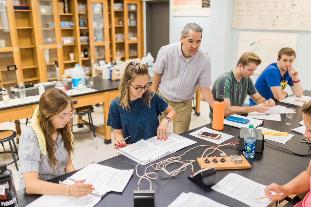 A group of students work on worksheets at a lab table, with a teacher supervising. Lab equipment, notebooks, and drinks are on the table. Shelves with supplies and posters are visible in the background.