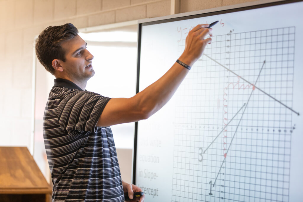 A person in a striped shirt writes a mathematical equation and draws lines on a large graph displayed on a whiteboard in a classroom, demonstrating effective teacher training techniques.