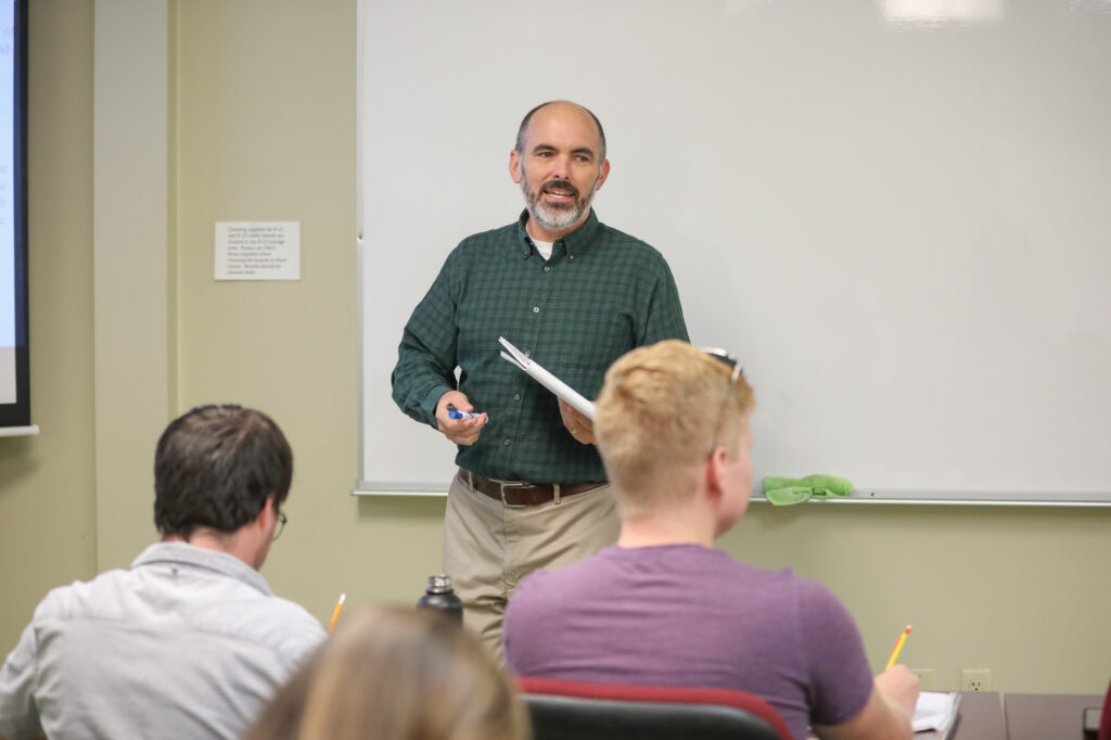 A man stands in front of a whiteboard holding papers and a marker, addressing three seated people who are facing him. The scene appears to be a classroom or lecture setting.