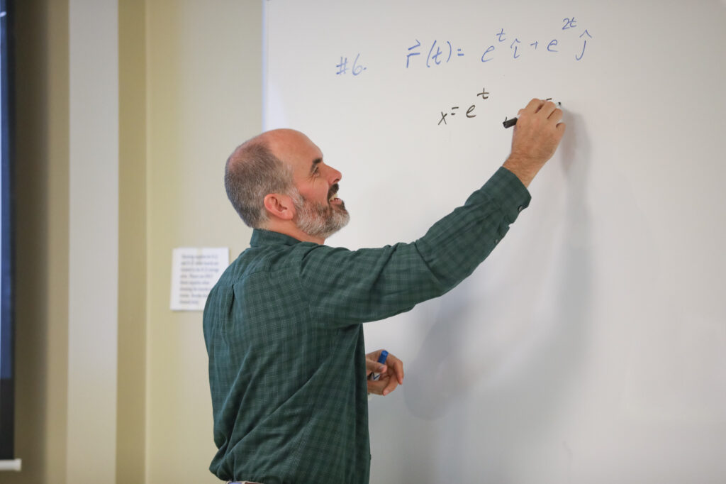 A man with a beard writes mathematical equations involving exponents on a whiteboard with a black marker. He is wearing a green plaid shirt and standing in a classroom.