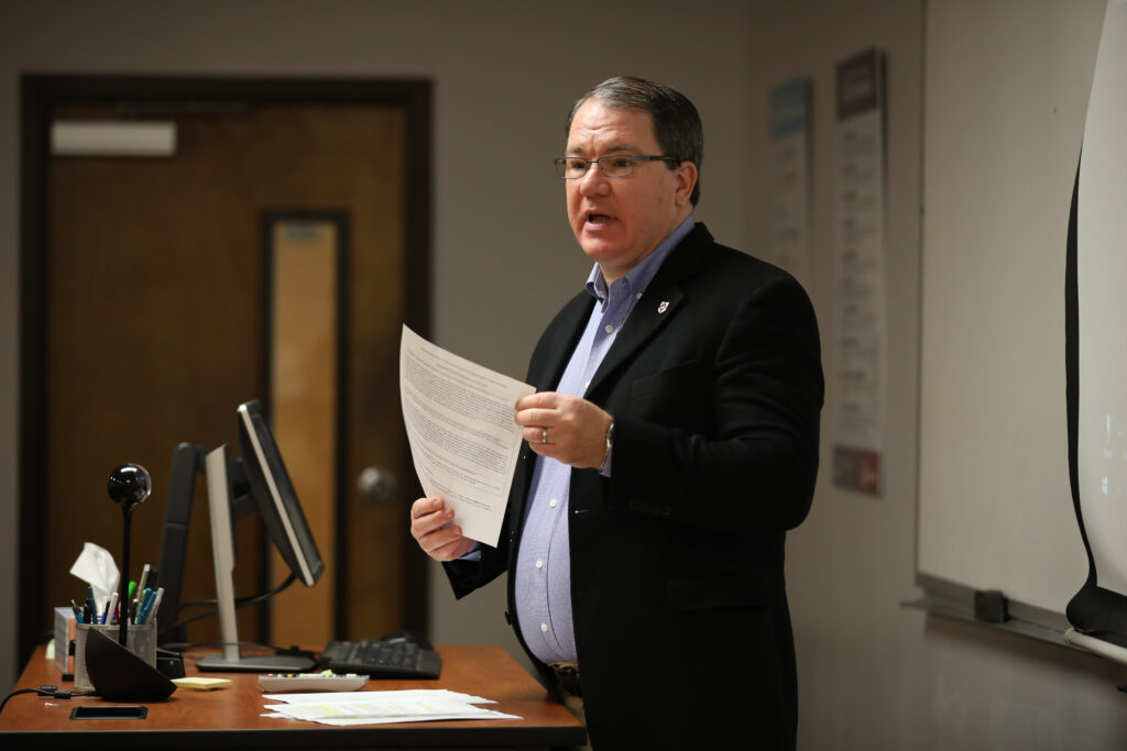 A man in a suit jacket is standing at a lectern in a classroom, holding papers and speaking. Emphasizing professional education, he is surrounded by a computer monitor, keyboard, office supplies, a whiteboard, and educational posters on the wall.