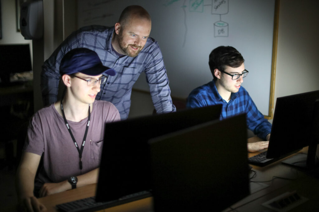 Three people are in a dimly lit room with computer monitors, collaborating on Cybersecurity Management. Two are seated and typing, while one stands observing their screens. A whiteboard with diagrams is visible in the background.