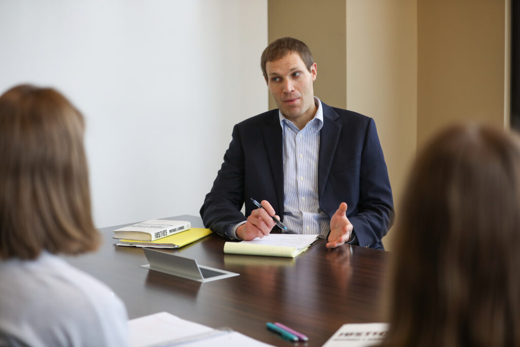 A man in a suit sits at a table speaking to two people. He has a notebook, pen, and folders in front of him. The two people are seen from behind. They are in a meeting or interview setting.