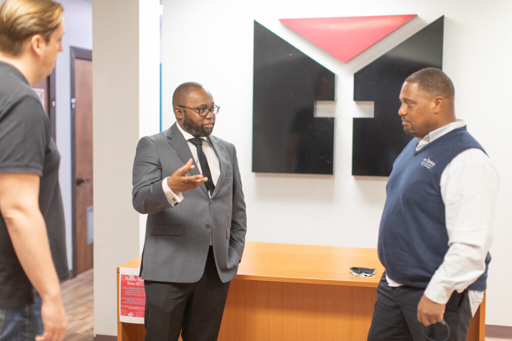 Three men are having a conversation in an office setting, embodying entrepreneurial leadership. One man in a suit gestures while talking. They stand near a wooden desk with glasses on it and a large geometric wall logo behind them.