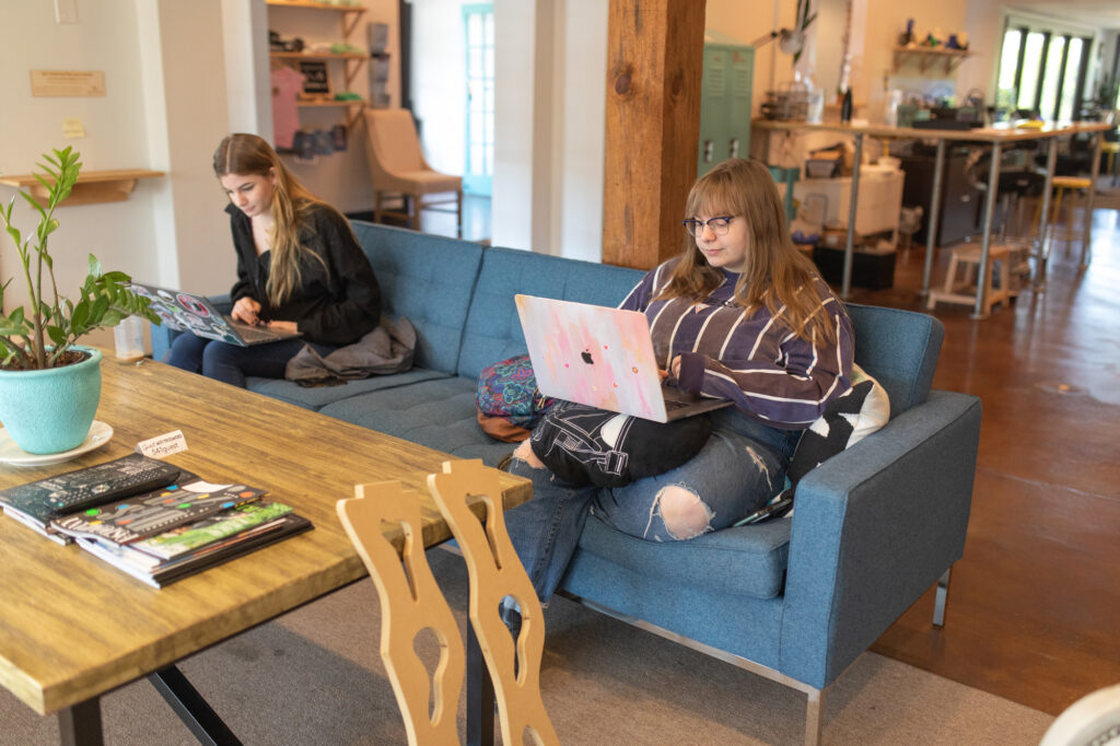 Two people sitting on a blue couch using laptops in a cozy, open space filled with books, plants, and hints of graphic design art. The background features shelves, lockers, and various creative items.
