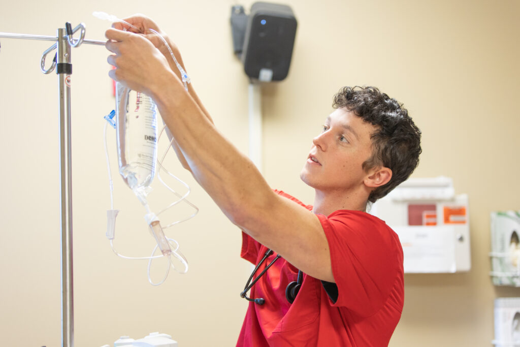 A person in a red shirt adjusts an IV bag on a metal stand in a medical setting in Hendersonville, with beige walls and office supplies visible in the background.