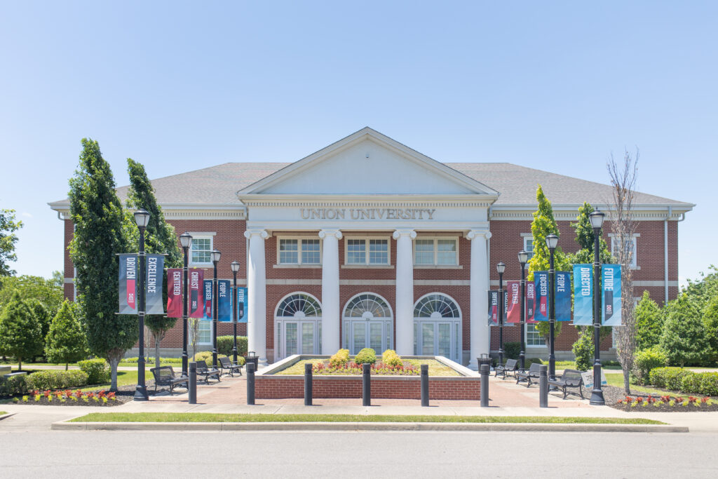 A large brick building with white columns and a sign reading Union University above the entrance, located near Hendersonville. Flags with academic program names hang from lampposts along a walkway lined with trimmed bushes and flowers.