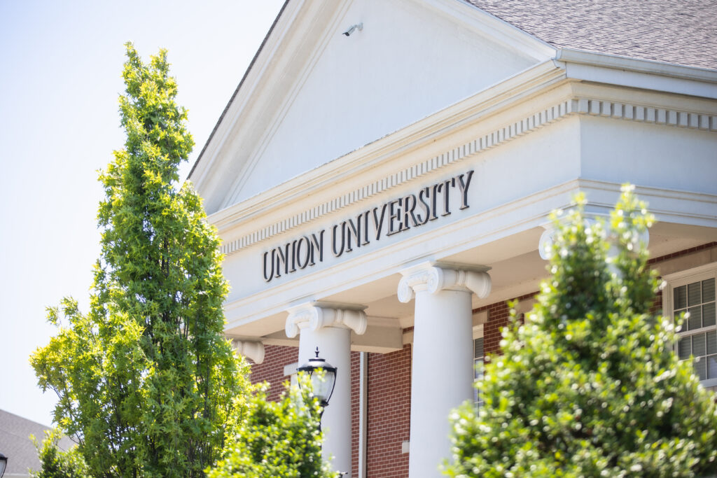 The front entrance of a building with white columns and Union University written above, partially framed by green trees under a clear Hendersonville sky.