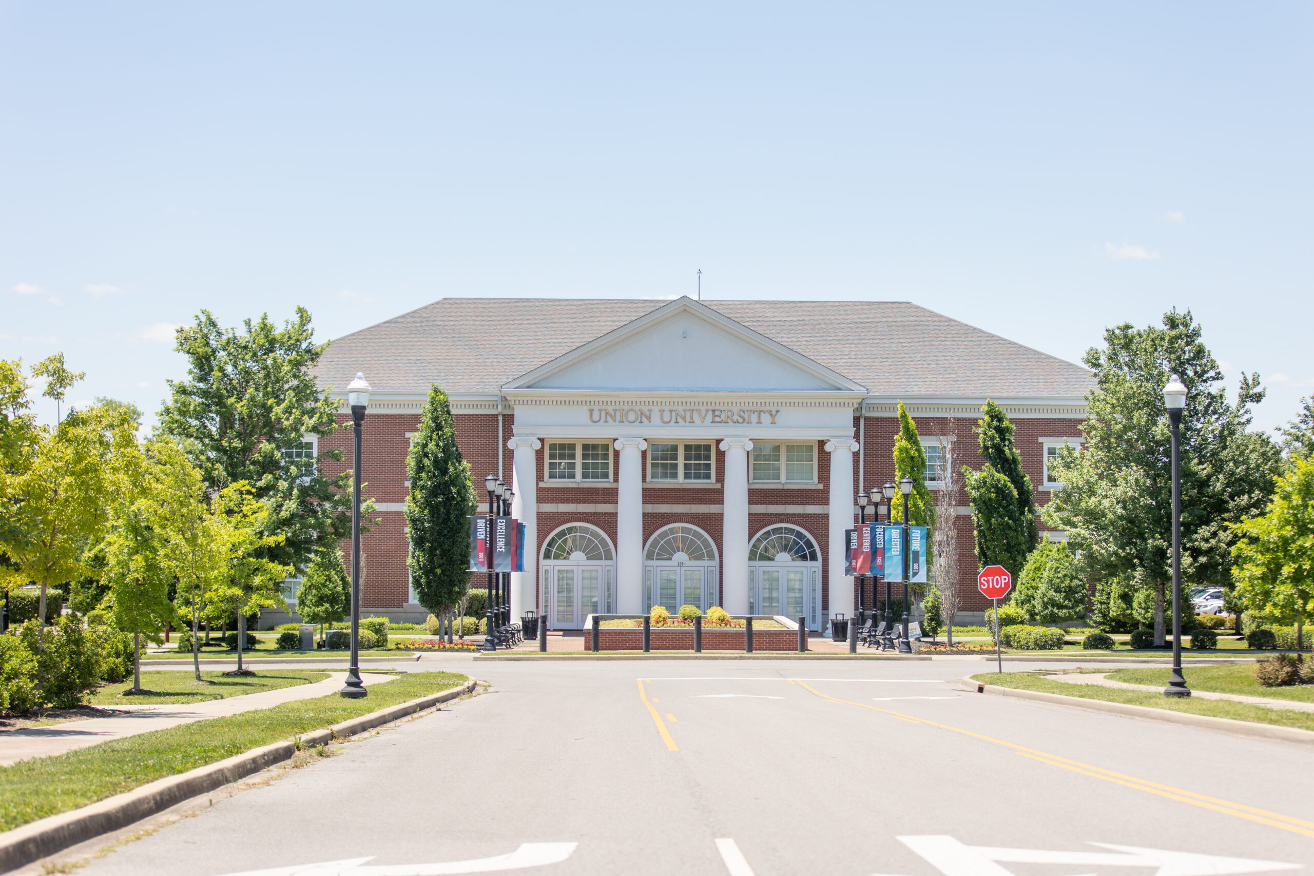 A large brick building with white columns and a sign reading Union University above the entrance, surrounded by trees and banners, sits near a stop sign at the corner of the road in Hendersonville, just outside Nashville.