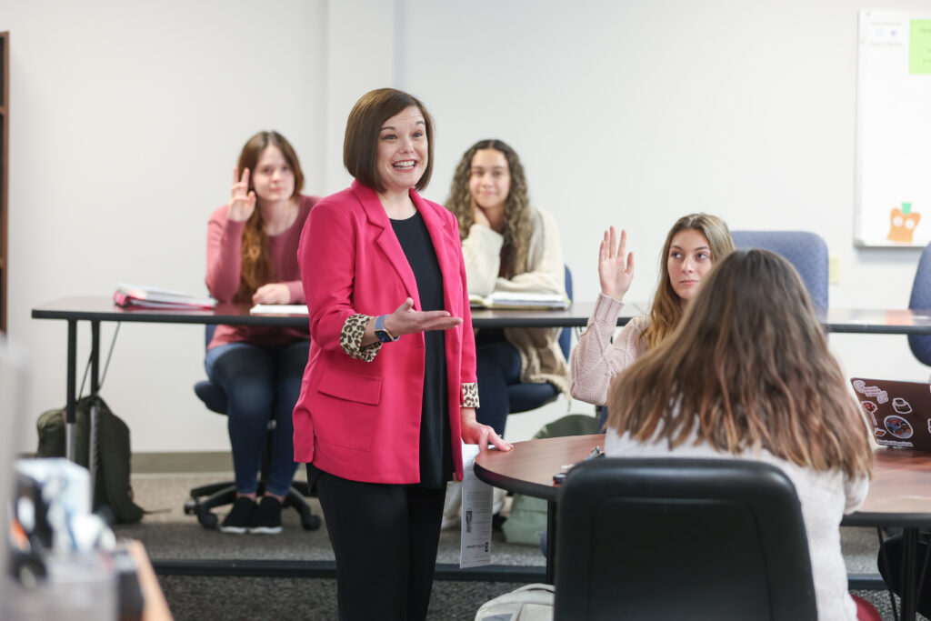 A woman in a pink blazer stands and speaks to a classroom of students enrolled in an MA in Education program, some raising their hands while others are seated with notebooks and laptops.