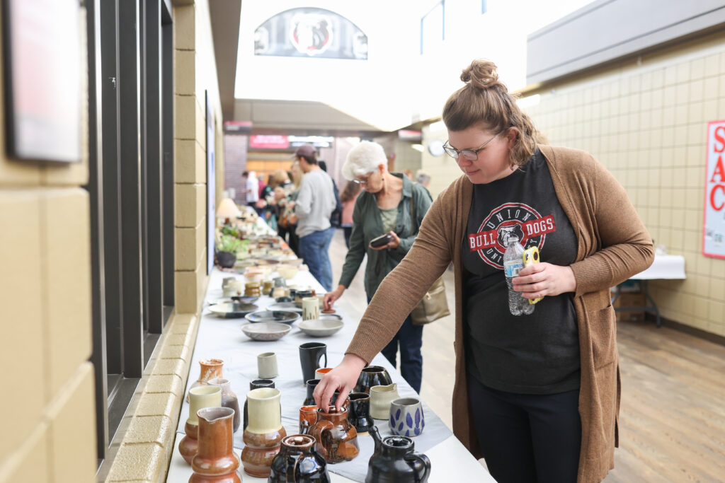 A woman wearing glasses and a Bulldogs t-shirt examines ceramic mugs and bowls on a table at an indoor market while other people browse in the background.