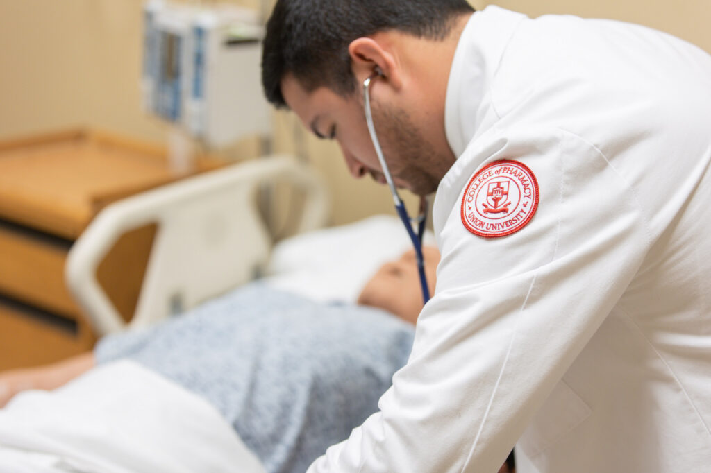 A medical professional with a Doctor of Pharmacy degree in a white coat uses a stethoscope to examine a patient lying in a hospital bed. The focus is on the PharmD, with the patient partially visible and out of focus.