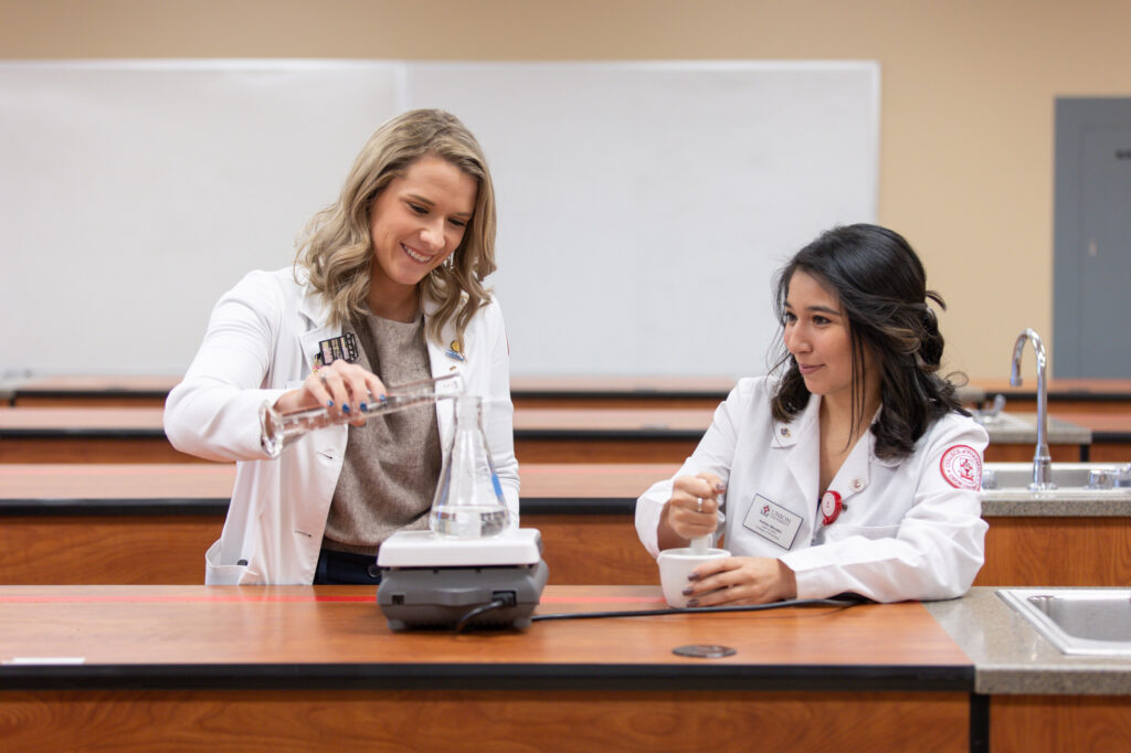 Two women in laboratory coats work at a lab station; one pours liquid into a flask on a hot plate while the other grinds a substance in a mortar, showcasing hands-on training essential for earning a PharmD or pharmacy degree in a classroom setting.
