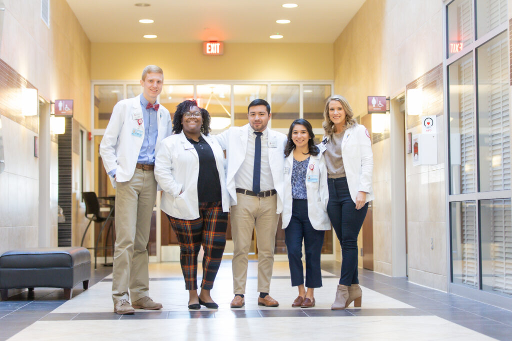 Five people in white lab coats stand together in a well-lit hallway. They are smiling and appear to be a diverse group of pharmacy professionals or students, possibly enrolled in a Two-Year Track program.