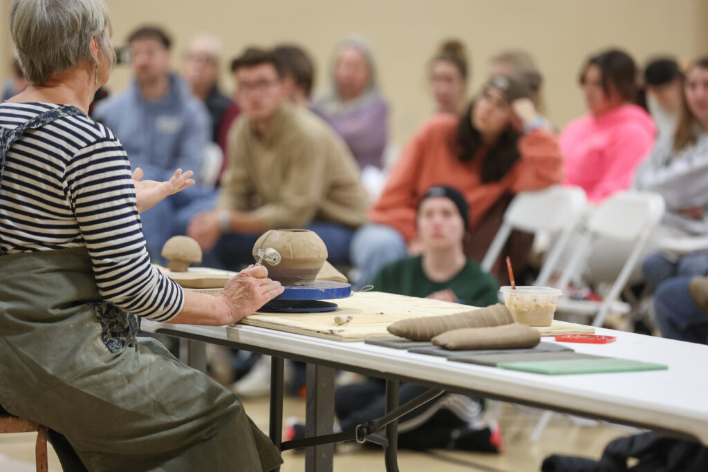 A person demonstrates ceramics techniques at a table with clay and tools, while a group of people seated in the background watch this art form attentively.