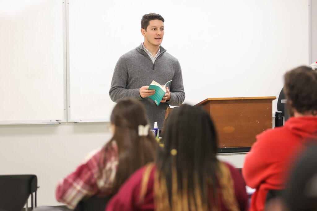 A man stands at a podium holding a book, speaking to a seated group of students in a classroom with a whiteboard in the background, as he discusses Ancient Languages.