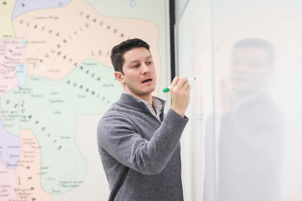 A man in a gray sweater writes on a whiteboard with a green marker, possibly discussing topics in Applied Linguistics. A colorful map with various regions and names is visible on the wall in the background.