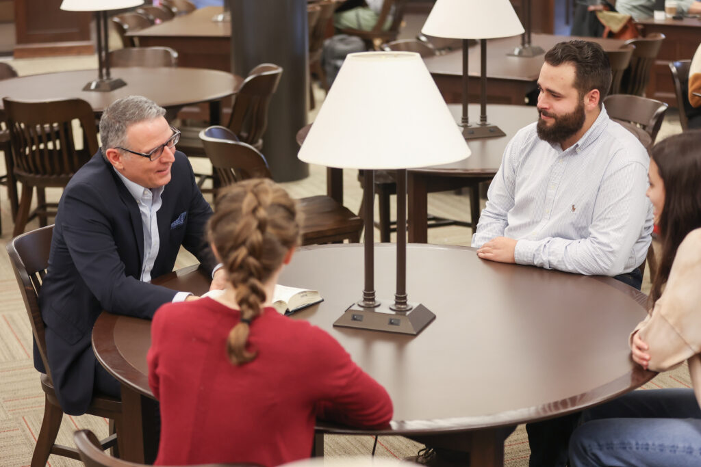 Four people sit around a round table in a library or study area, engaged in conversation. A lamp is on the table, and books and chairs are visible in the background.