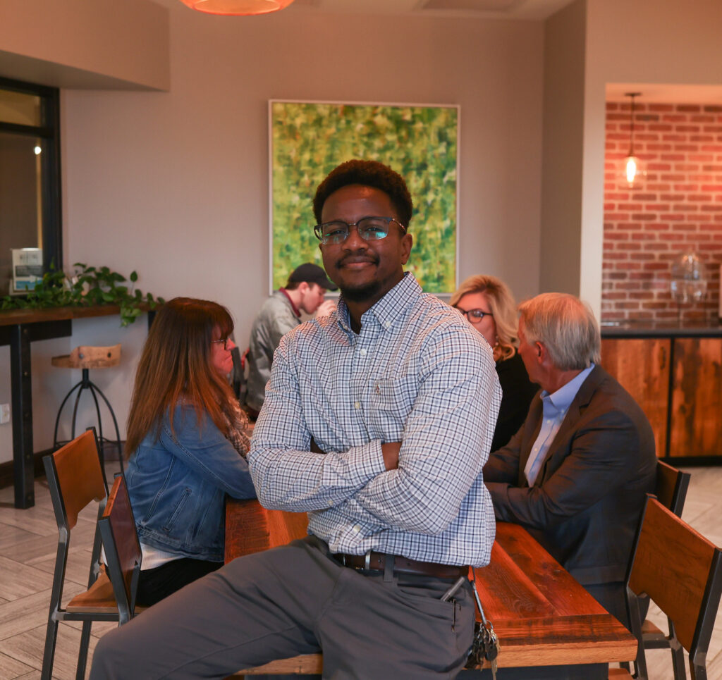 A man in glasses and a checked shirt sits on the edge of a wooden table, arms crossed, in a modern room. Four people—perhaps discussing a Professional Studies or Science Degree—are at the table. A green abstract painting hangs behind them.