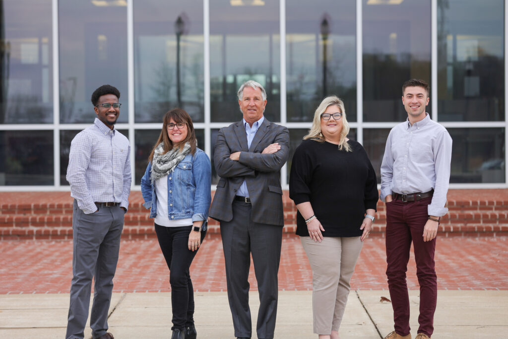 Five people stand outdoors in front of a building with large windows. The group, representing strong organizational leadership within ASPS, includes two men and three women in business casual attire, posing for a group photo.