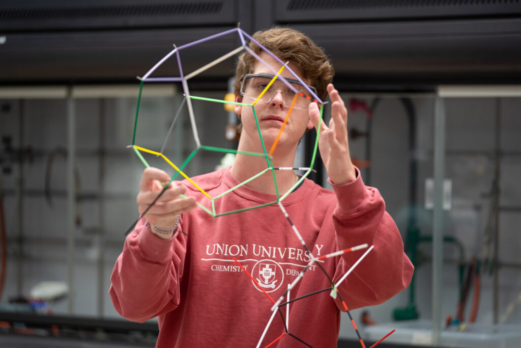 A person wearing safety glasses and a red Union University Chemistry Department sweatshirt examines a colorful geometric structure made of connected sticks in a science lab.