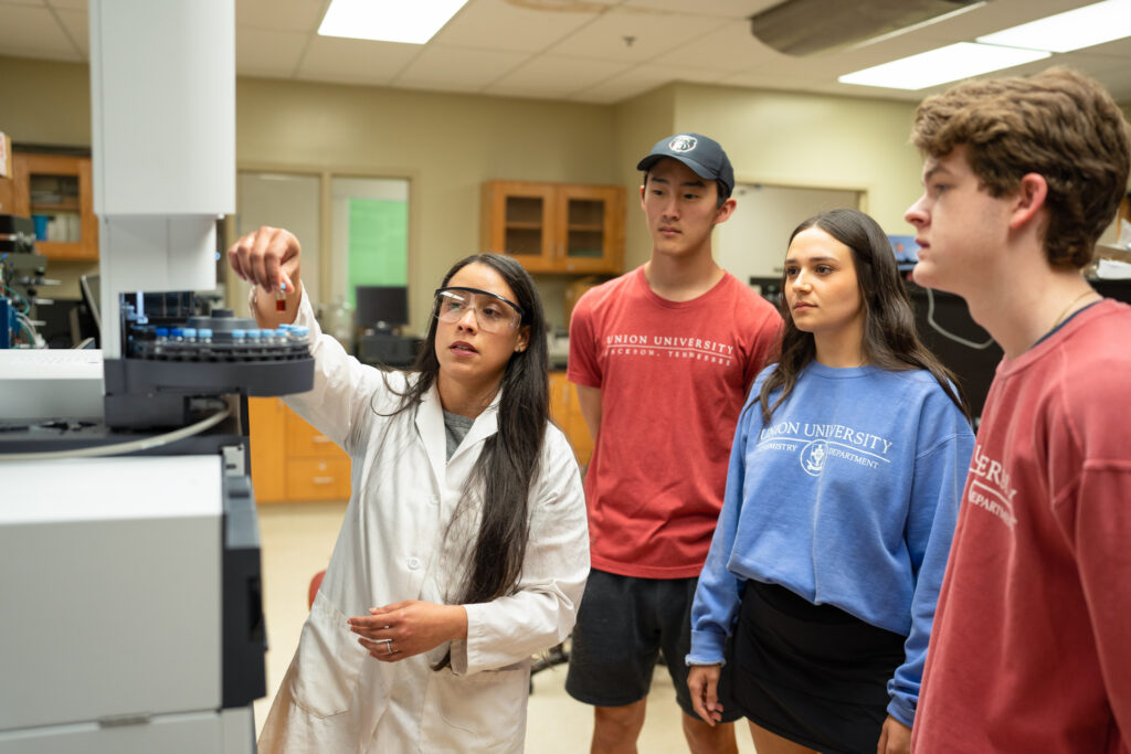 A woman in a lab coat and safety glasses demonstrates chemistry equipment to three students in casual clothing inside a laboratory classroom.