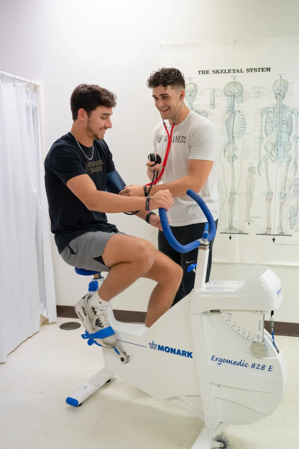 A person pedals a stationary exercise bike while another person in a t-shirt and holding a stethoscope measures their blood pressure in a medical room with a skeletal system chart on the wall.