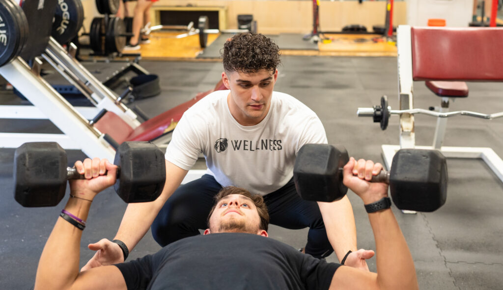 A man is bench pressing dumbbells while lying on a bench. Another man in a white Wellness shirt, likely trained in Exercise Science, stands behind him, spotting and assisting in a gym setting.