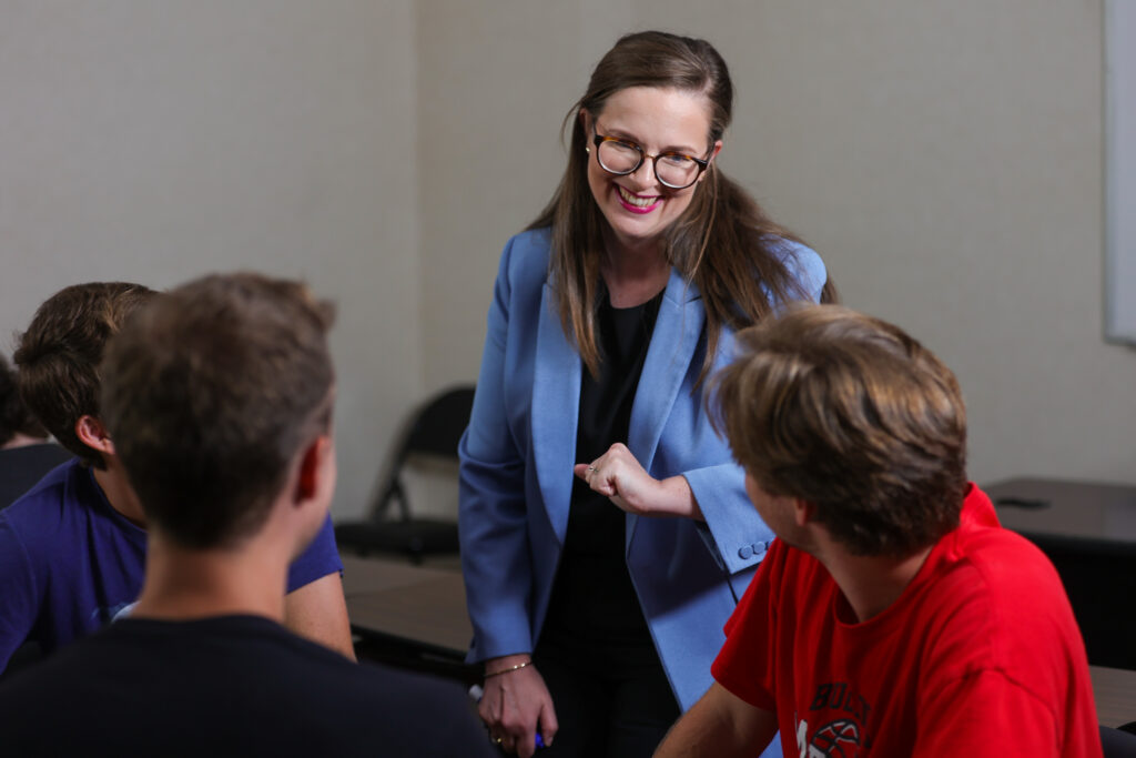 A woman in a blue blazer stands and smiles while talking to three seated people in a classroom or meeting room, leading an engaging discussion about Public Relations.