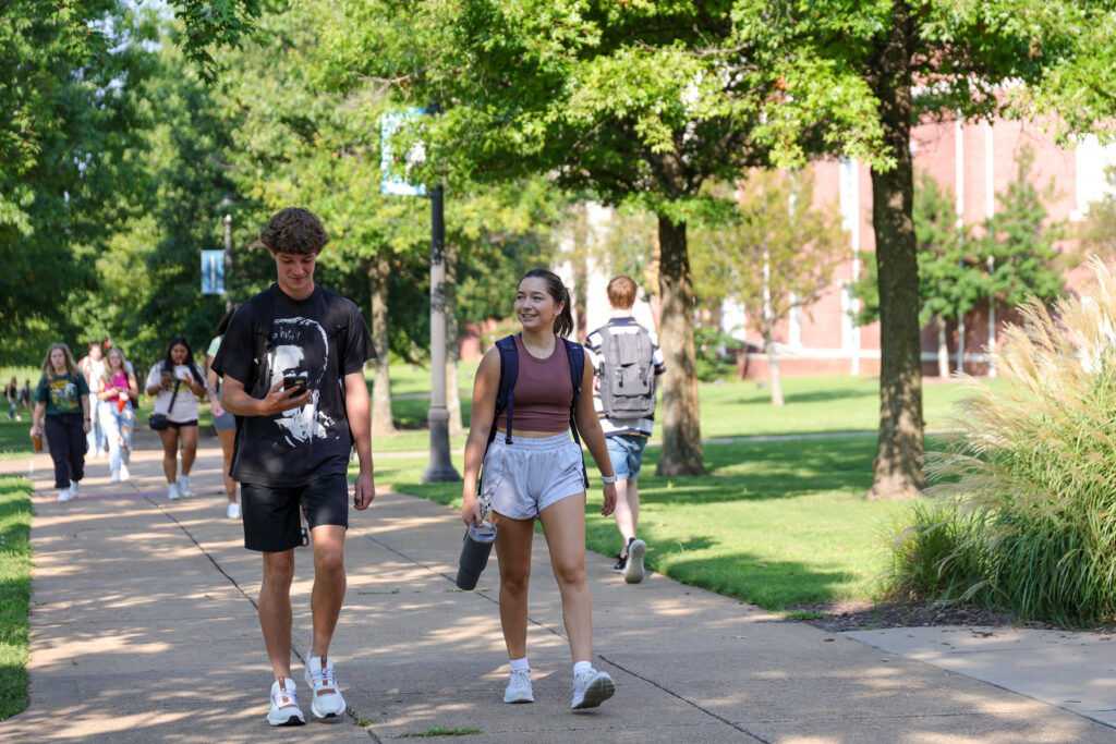Students walk along a tree-lined sidewalk on Union's main campus in Jackson, Tennessee, enjoying a sunny day. In the foreground, two students talk and smile with backpacks and water bottles while others chat in the background.