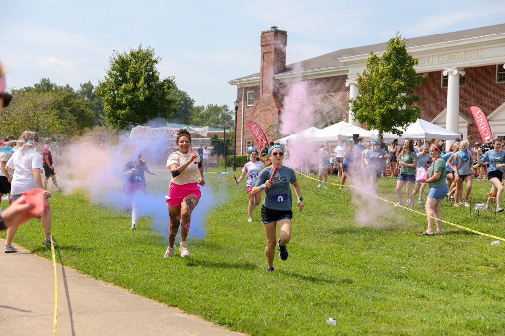 A group of people run on a grassy lawn through clouds of colored powder during an outdoor event, while onlookers stand nearby. A brick building and several tents are in the background.