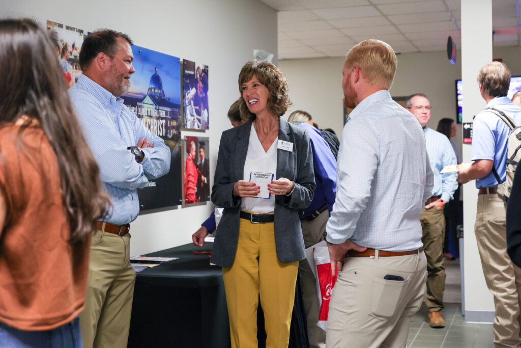 A group of people converse in a hallway near a table with pamphlets. One woman in a blazer and yellow pants, possibly discussing Business Administration, smiles as she talks with two men in business-casual attire. Posters are visible on the wall.