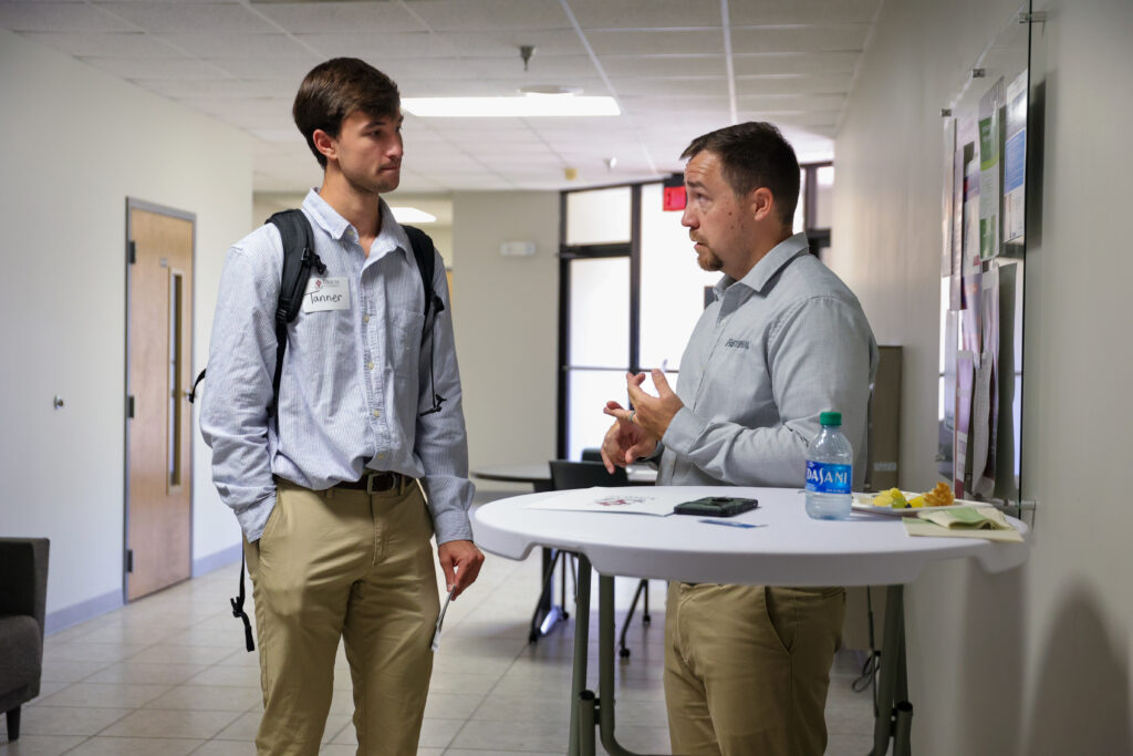 Two men in business administration attire—light blue shirts and khaki pants—stand at a round table in a hallway, discussing finance. One has a backpack; the table holds a water bottle, papers, and a plate with food.