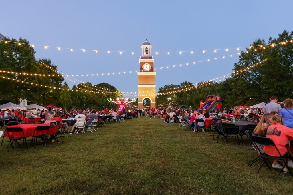 People gather at tables on a grassy lawn for an outdoor event in the evening at Tennessee’s Union's main campus in Jackson, with string lights overhead, a tall brick clock tower in the background, and inflatable attractions to the right.
