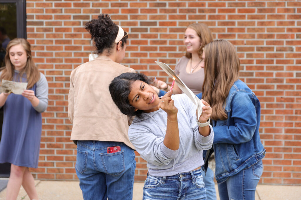 A group of five young women are standing outside near a brick wall, holding and examining art projects. One woman in the foreground smiles at the camera while proudly showing off her drawing.