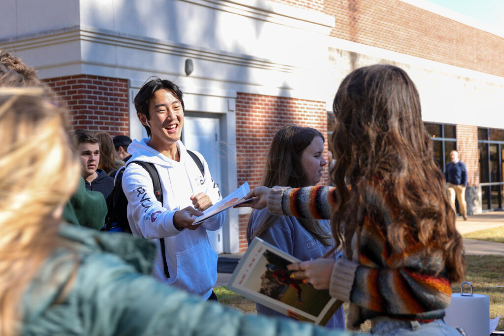 A group of students stand outside a brick building. One student in a white hoodie smiles as he takes a paper from a student in a striped sweater. Others hold folders and talk nearby.