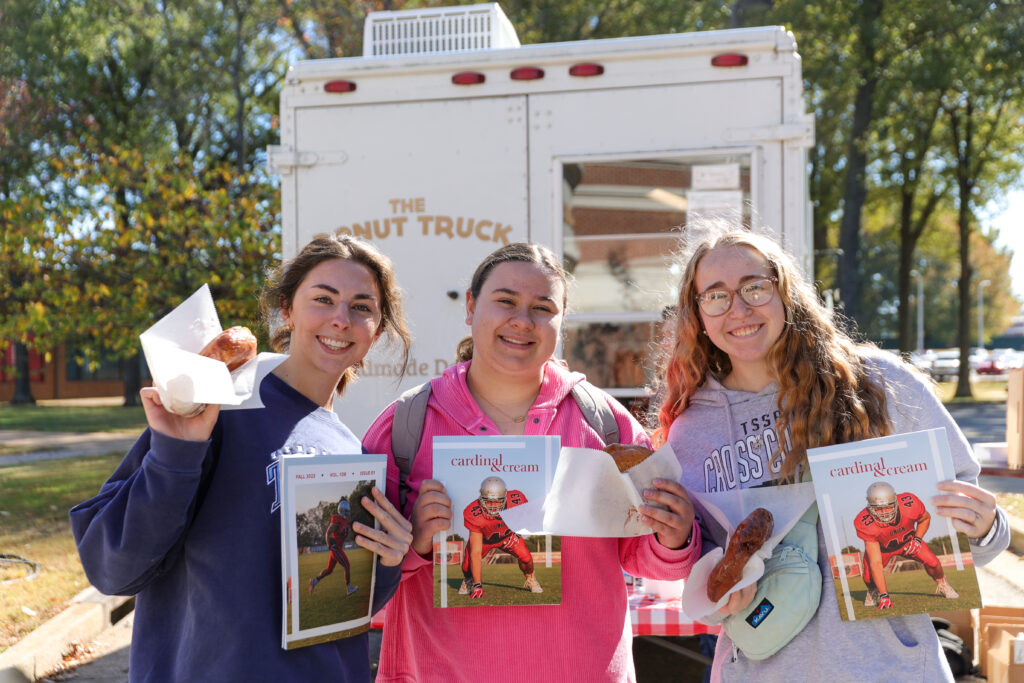 Three young women stand outdoors in front of a white food truck on Union's main campus in Jackson, each holding a pastry and a copy of “carting cream” magazine. Sunlight filters through green trees, capturing a vibrant Tennessee day.