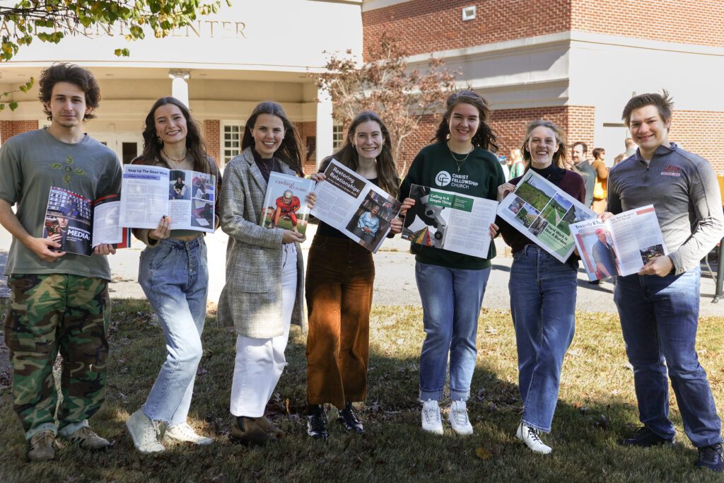 Seven young adults stand outside a building, holding open magazines or brochures and smiling at the camera. Dressed in casual clothing, they enjoy the sunny day—showcasing their passion for storytelling and Digital Media Communications.