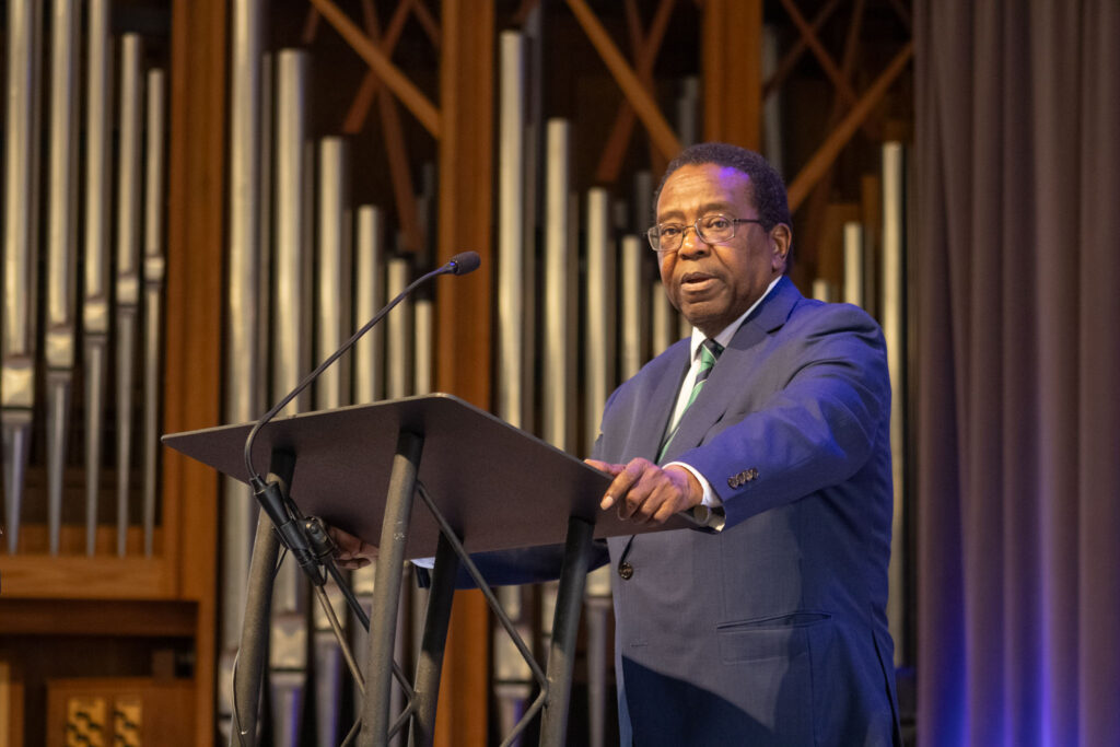 A man in a dark suit stands at a podium with microphones, speaking. Behind him are large organ pipes and wooden paneling. A gray curtain is visible on the right side of the image.