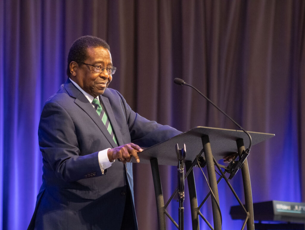 A man in a dark suit, striped tie, and glasses stands at a podium, speaking into a microphone about Theology. A keyboard is visible on the right, and a curtain with blue lighting is in the background.